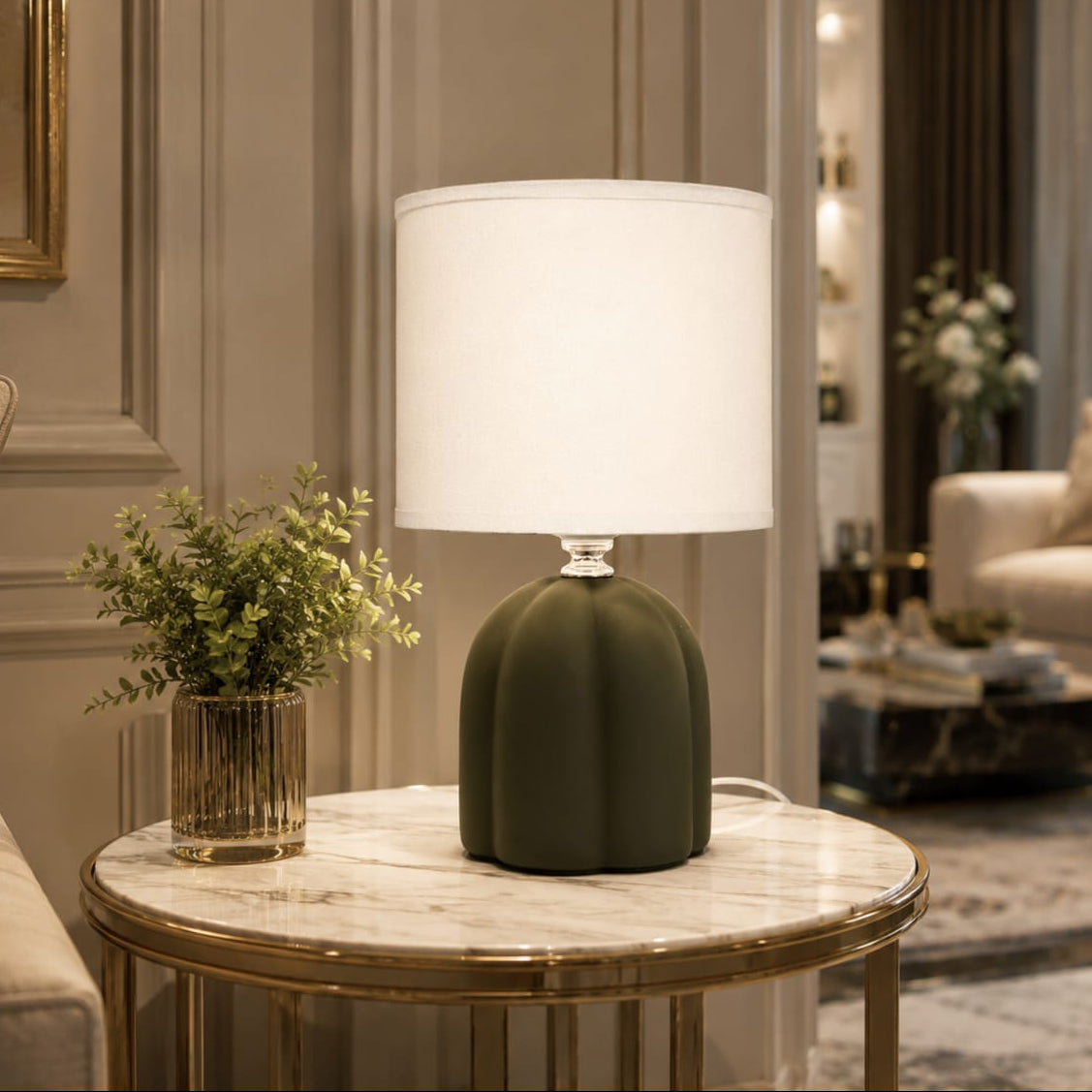 A modern table lamp with a ribbed dark olive green base and a white drum shade, placed on a marble-topped side table in a stylishly decorated living room.