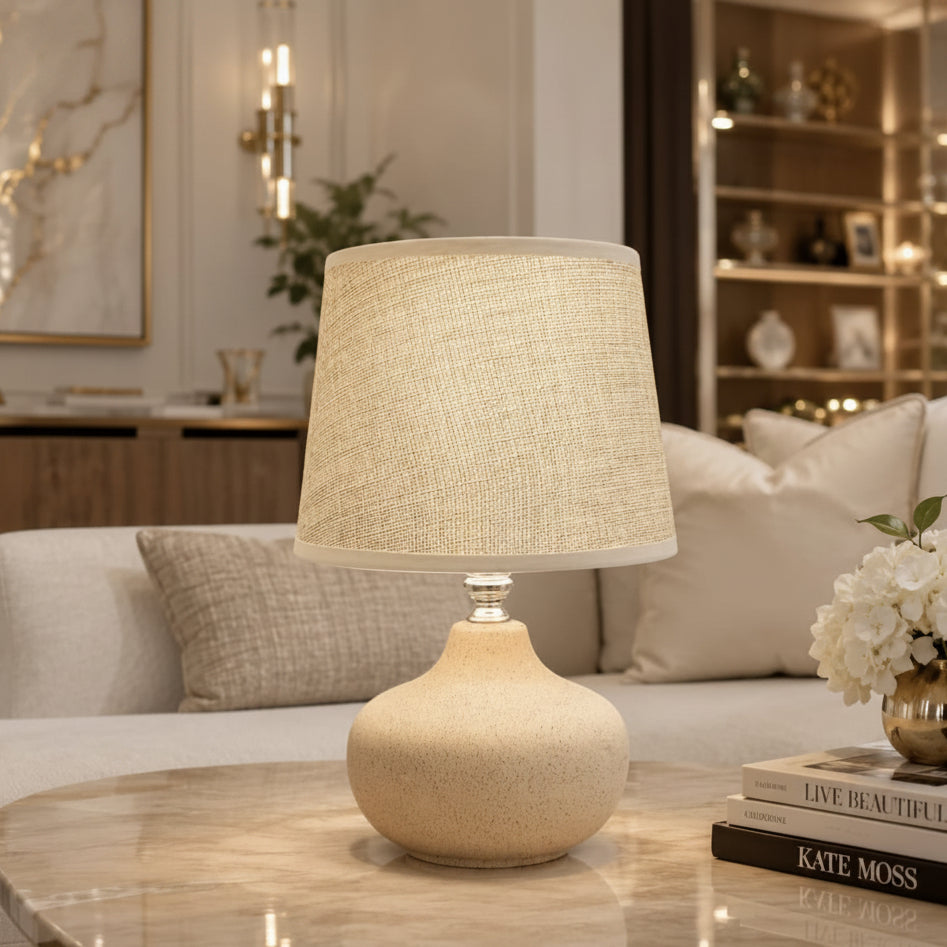 A table lamp with a textured beige fabric drum shade and a matching speckled beige ceramic base, shown lit on a marble-top table in a modern living room setting.