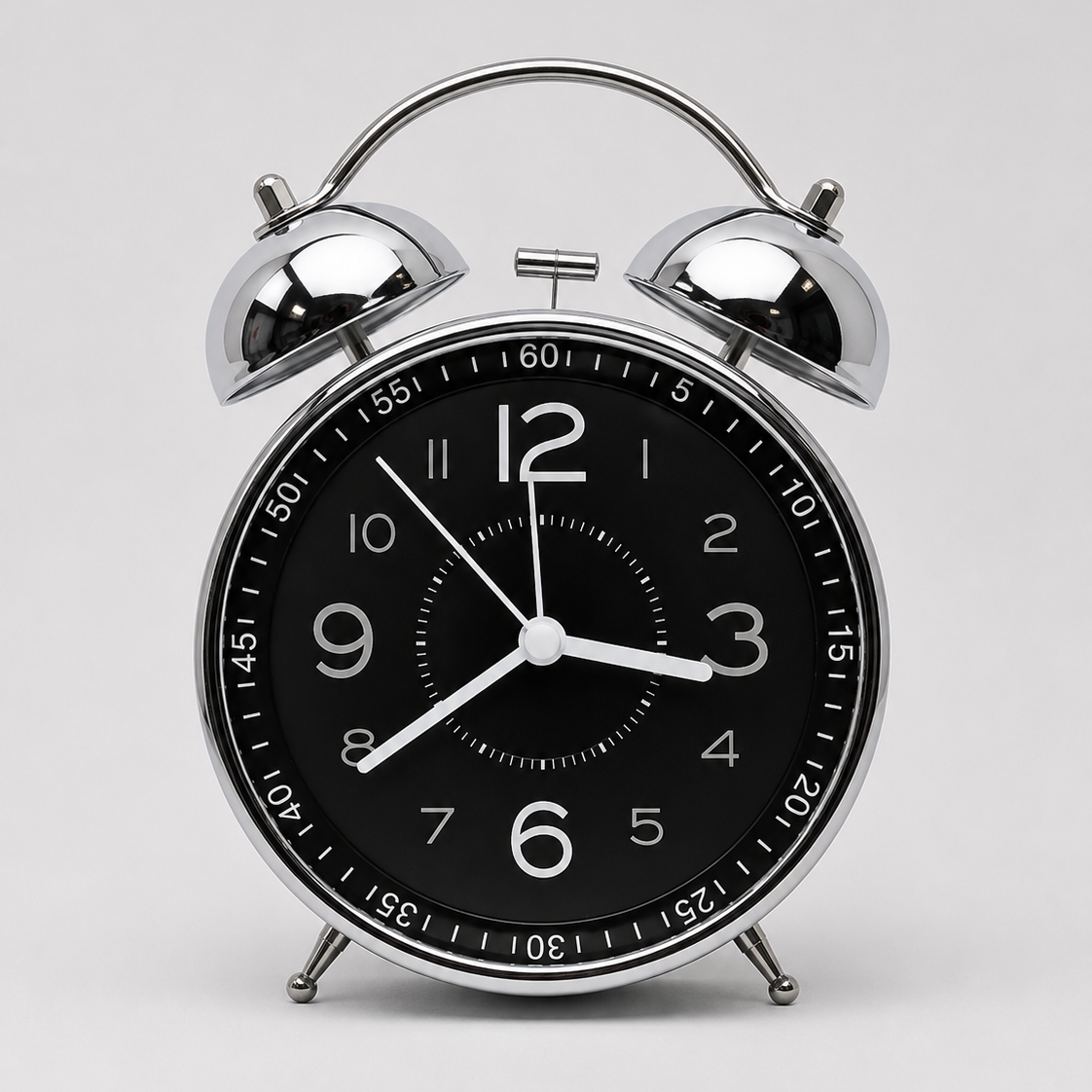 A classic twin-bell alarm clock with a polished silver metal body and a black face, featuring white Arabic numerals and hands, set against a light gray background.